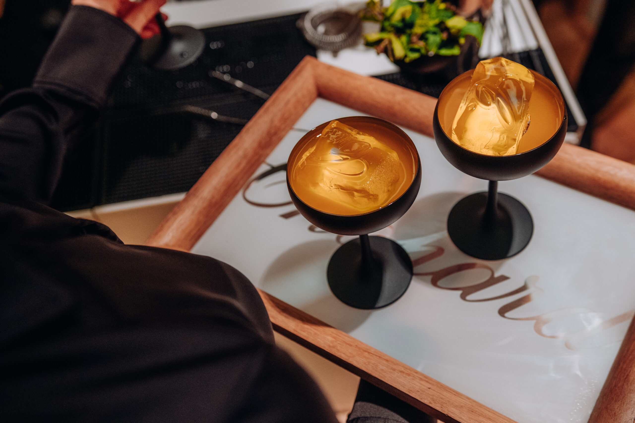 Zwei goldene Cocktailgläser auf schwarzem Stiel stehen auf weißem Tablett, gehalten von Person in schwarzer Kleidung