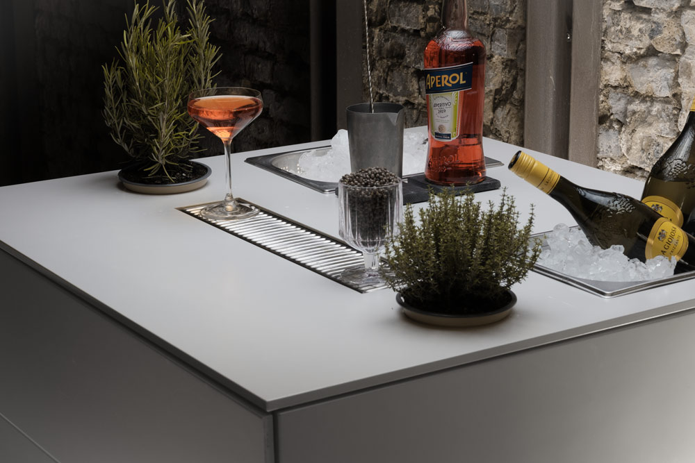 Mobile Bar mit Cocktailglas, Aperol-Flasche, Champagner im Eiskübel und Kräuterdekoration auf weißer Arbeitsplatte