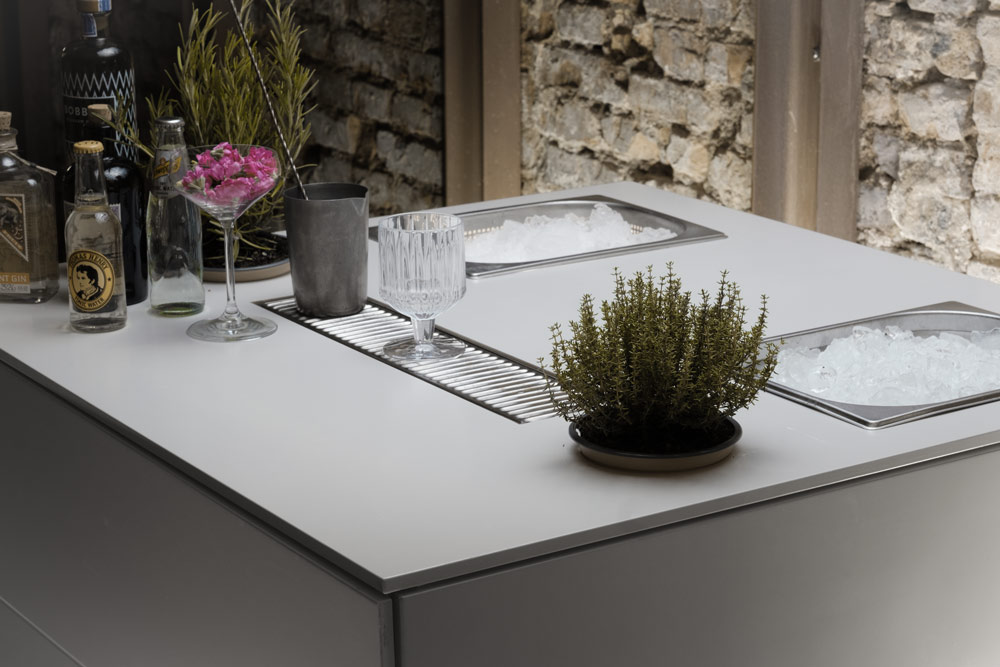 Moderne mobile Bar mit weißer Arbeitsplatte, Spülbecken, Cocktailzubehör und Kräuterpflanze vor Steinwand