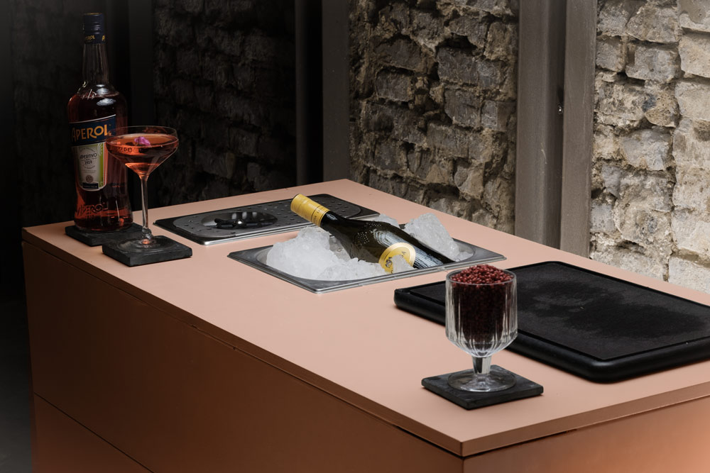 Elegante mobile Bar mit Aperol-Flasche, Cocktailgläsern und gekühlter Champagnerflasche auf Eis vor rustikaler Steinwand