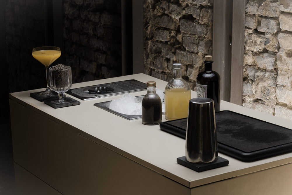 Moderne mobile Bar mit Cocktailgläsern, Flaschen und Barzubehör vor rustikaler Steinwand in elegantem Ambiente