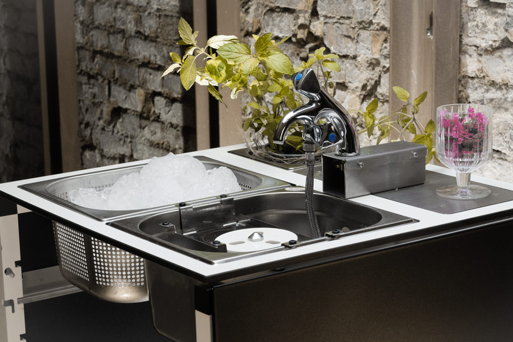 Mobile Cocktailbar mit Edelstahl-Spüle, Wasserhahn und Weinglas vor rustikaler Steinwand mit grünen Pflanzen